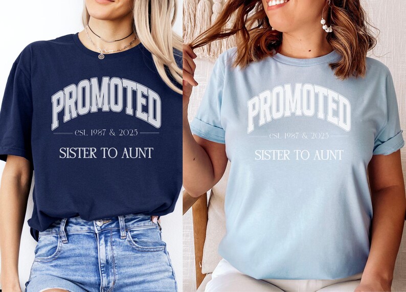 May include: Two t-shirts, one navy blue and one light blue, each with the word "PROMOTED" in a white, arched font. Below, the text reads "est. 1987 & 2025" and "SISTER TO AUNT". The shirts are casual, with rolled sleeves.