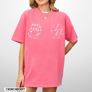 May include: A pink t-shirt with white dice graphics, each featuring heart-shaped pips. The text "Bailyn's Bach" and "Las Vegas 2024" are printed on the shirt. The shirt is a loose fit and has short sleeves.