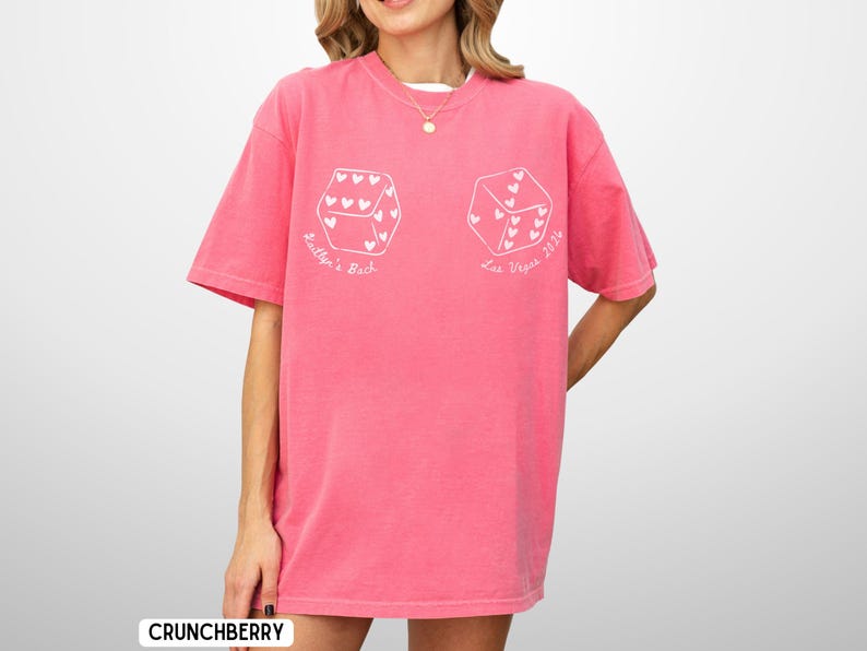 May include: A pink t-shirt with white dice graphics, each featuring heart-shaped pips. The text "Bailyn's Bach" and "Las Vegas 2024" are printed on the shirt. The shirt is a loose fit and has short sleeves.