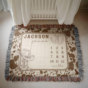 May include: A rectangular woven throw blanket featuring a brown and white cowhide print border and a central cream-colored panel. The design includes the name Jackson James, a cowboy hat, a pair of horseshoes, and numbers 1 through 12.