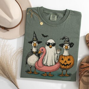 May include: Moss green t-shirt featuring a Halloween-themed graphic of three geese dressed in costumes. One goose wears a witch hat and cape, another is a ghost with sunglasses, and the third is a jack-o'-lantern. A pink flamingo floatie is also present.