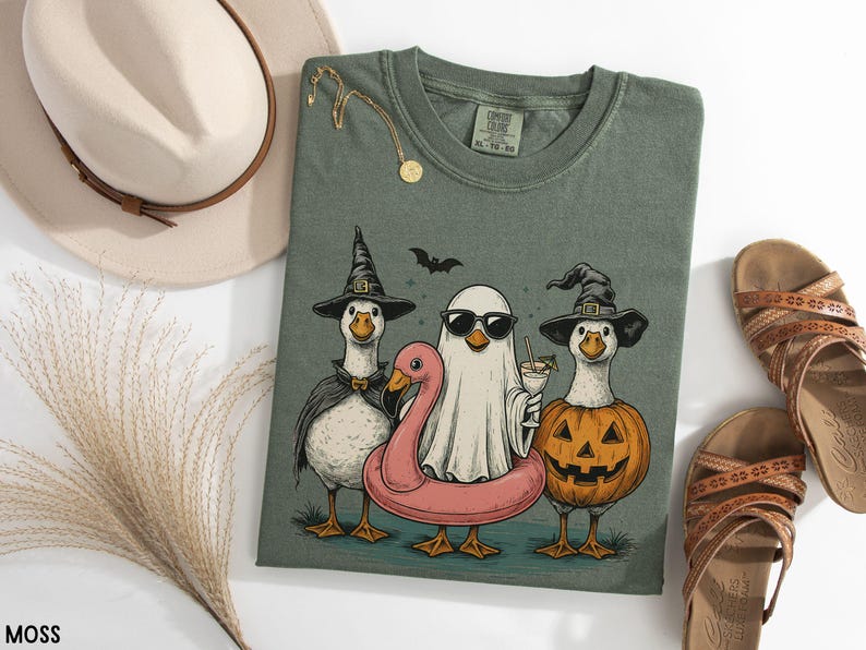 May include: Moss green t-shirt featuring a Halloween-themed graphic of three geese dressed in costumes. One goose wears a witch hat and cape, another is a ghost with sunglasses, and the third is a jack-o'-lantern. A pink flamingo floatie is also present.