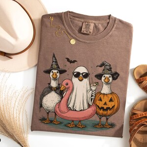 May include: Espresso-colored t-shirt featuring a Halloween-themed graphic. The design includes three geese dressed as a witch, a ghost, and a jack-o'-lantern, with a pink flamingo floatie. A bat flies above them. A gold necklace and sandals are also visible.