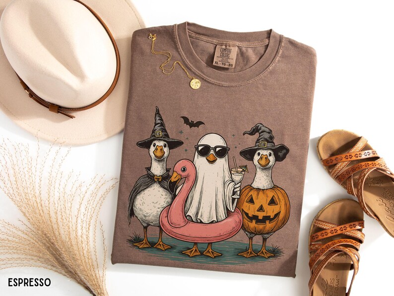 May include: Espresso-colored t-shirt featuring a Halloween-themed graphic. The design includes three geese dressed as a witch, a ghost, and a jack-o'-lantern, with a pink flamingo floatie. A bat flies above them. A gold necklace and sandals are also visible.
