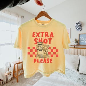 K&ouml;nnte beinhalten: Gelbes T-Shirt mit dem roten Schriftzug "EXTRA SHOT PLEASE". Darunter befindet sich eine Grafik einer Kaffeemaschine, die von einem rot-orangen Karomuster umrahmt ist.