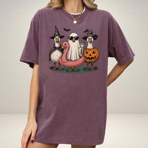 May include: A purple t-shirt featuring a Halloween-themed graphic. The graphic depicts three cartoon geese dressed in costumes: a witch, a ghost, and a jack-o'-lantern. The ghost is wearing sunglasses and the geese are standing on a pink inflatable flamingo.