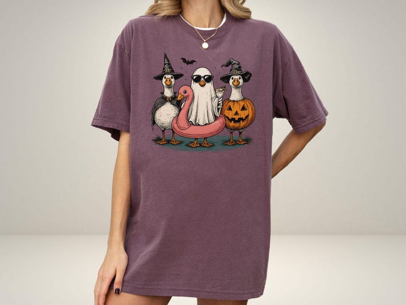 May include: A purple t-shirt featuring a Halloween-themed graphic. The graphic depicts three cartoon geese dressed in costumes: a witch, a ghost, and a jack-o'-lantern. The ghost is wearing sunglasses and the geese are standing on a pink inflatable flamingo.