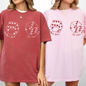 May include: Two oversized t-shirts, one brick red and one light pink. Each shirt features a dice graphic with heart-shaped pips and the text "Kaitlyn's Bach Las Vegas 2024". The shirts have short sleeves and a relaxed fit.