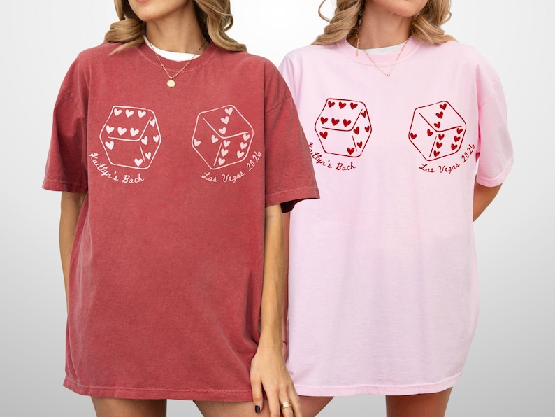 May include: Two oversized t-shirts, one brick red and one light pink. Each shirt features a dice graphic with heart-shaped pips and the text "Kaitlyn's Bach Las Vegas 2024". The shirts have short sleeves and a relaxed fit.