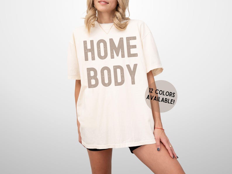 May include: A white t-shirt with the text "HOME BODY" printed in brown on the front. The text has a distressed look. The t-shirt is being modeled by a person. 12 colors are available.