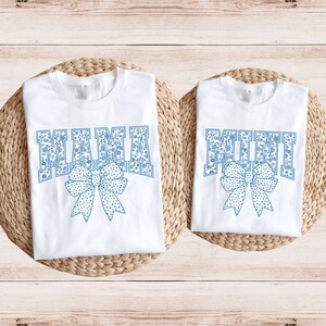 May include: Two white t-shirts with blue floral print lettering. The larger shirt says "MAMA" and the smaller shirt says "MINI". Both shirts feature a blue bow with white polka dots.