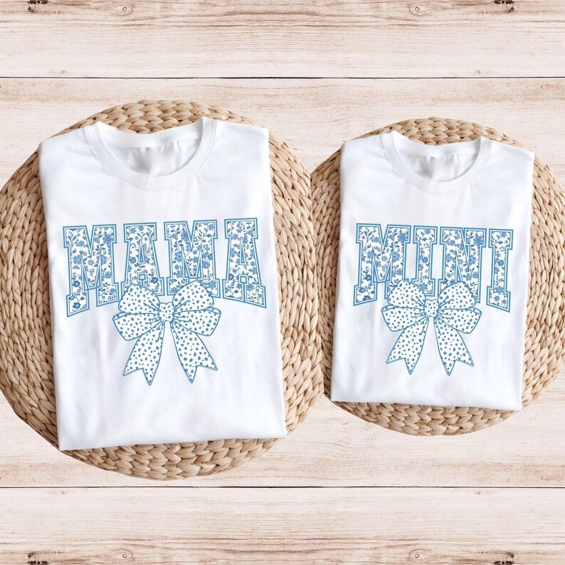 May include: Two white t-shirts with blue floral print lettering. The larger shirt says "MAMA" and the smaller shirt says "MINI". Both shirts feature a blue bow with white polka dots.
