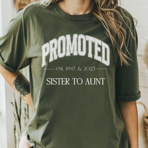 May include: Olive green t-shirt with the word "PROMOTED" in white, arched across the chest. Below, the text reads "est. 1987 & 2025" and "SISTER TO AUNT". The shirt has rolled-up sleeves.