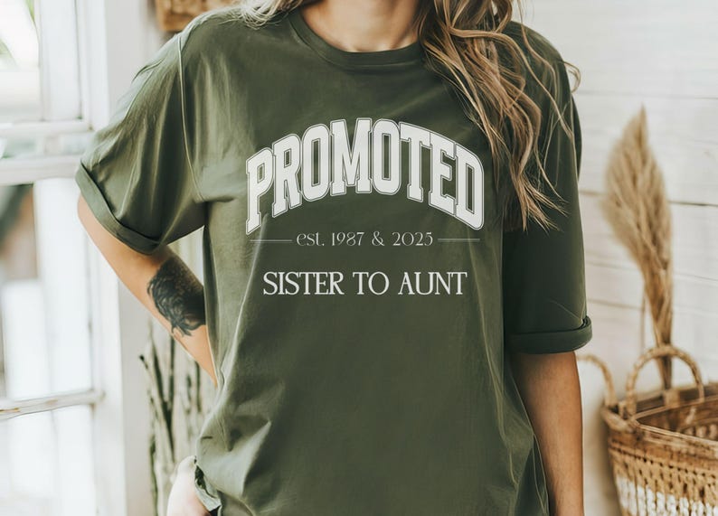 May include: Olive green t-shirt with the word "PROMOTED" in white, arched across the chest. Below, the text reads "est. 1987 & 2025" and "SISTER TO AUNT". The shirt has rolled-up sleeves.