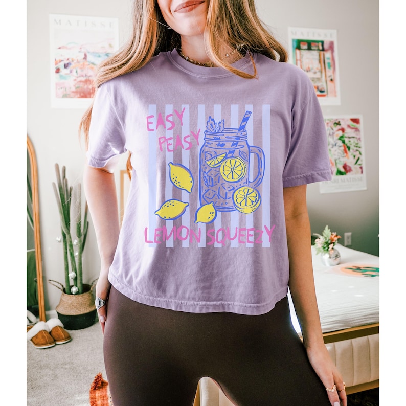 Pu&ograve; includere: Maglietta lilla con grafica a tema limonata. Il design include un barattolo con limoni e ghiaccio, e la scritta "Easy Peasy Lemon Squeezy". La maglietta ha maniche corte ed &egrave; di colore lilla chiaro.