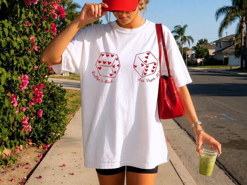May include: White t-shirt with red dice graphics, each with heart-shaped pips. Text on the shirt reads "Kaitlyn's Bach" and "Las Vegas 2021". A red purse and cap complement the shirt. The person is holding a green drink.