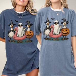 May include: Two blue t-shirts featuring a Halloween-themed graphic. The graphic depicts three cartoon geese dressed in Halloween costumes: a witch, a ghost, and a jack-o'-lantern. A pink flamingo floatie is also included.