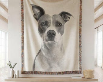 May include: A woven throw blanket featuring a grayscale portrait of a dog. The dog has a white and gray coat with attentive eyes. The blanket has a fringed border with a mix of colors and is displayed on a wall.