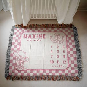 May include: A woven throw blanket with a pink and white checkered border and fringe edges. The center features the name Maxine, a cowboy hat, a pair of cowboy boots, a horseshoe, and the numbers 1 through 12 with the word months printed.