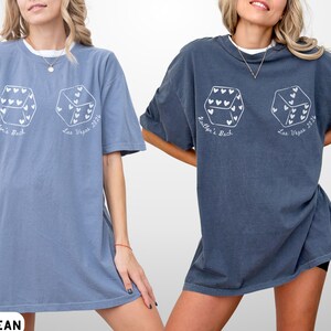 May include: Two oversized t-shirts, one blue jean and one denim, featuring a dice graphic with heart-shaped pips. The text "Kaitlyn's Back" and "Las Vegas 2026" are printed on the shirts. The shirts are worn by a person.