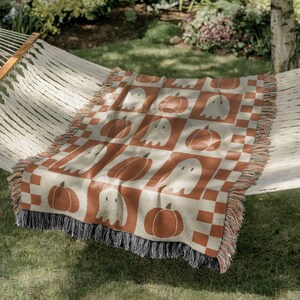 May include: A woven throw blanket with a Halloween theme. The blanket features a repeating pattern of pumpkins and ghosts in orange and off-white squares. The blanket has a fringed edge and is draped over a hammock.