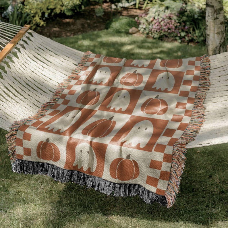 May include: A woven throw blanket with a Halloween theme. The blanket features a repeating pattern of pumpkins and ghosts in orange and off-white squares. The blanket has a fringed edge and is draped over a hammock.