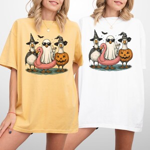 May include: Yellow and white t-shirts featuring a Halloween-themed graphic. The graphic includes cartoon ducks dressed as a witch, a ghost, and a jack-o'-lantern, with a pink flamingo floatie. Bats are flying above the characters.