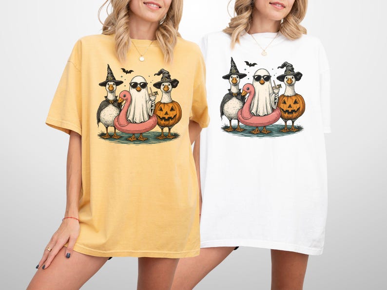 May include: Yellow and white t-shirts featuring a Halloween-themed graphic. The graphic includes cartoon ducks dressed as a witch, a ghost, and a jack-o'-lantern, with a pink flamingo floatie. Bats are flying above the characters.