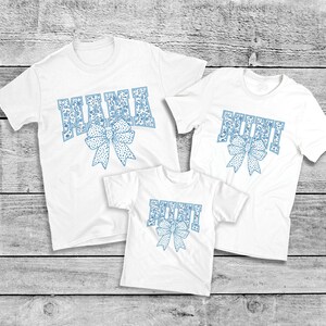 May include: Three white t-shirts with blue patterned text. The largest shirt says "MAMA" with a blue bow. The medium shirt says "MIMI" with a blue bow. The smallest shirt says "MINI" with a blue bow.