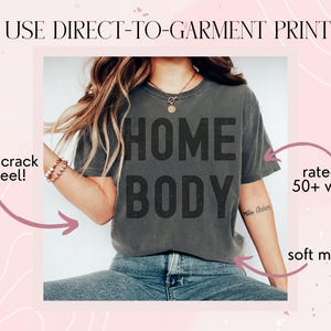 May include: A gray t-shirt with the text "HOME BODY" printed on it. The shirt is being worn by a person and is shown against a pink background with white swirls. The text "WE USE DIRECT-TO-GARMENT PRINTING" is at the top of the image. The text "won't crack or peel!" is on the left side of the image. The text "rated for 50+ washes" is on the right side of the image. The text "soft material" is at the bottom right of the image.