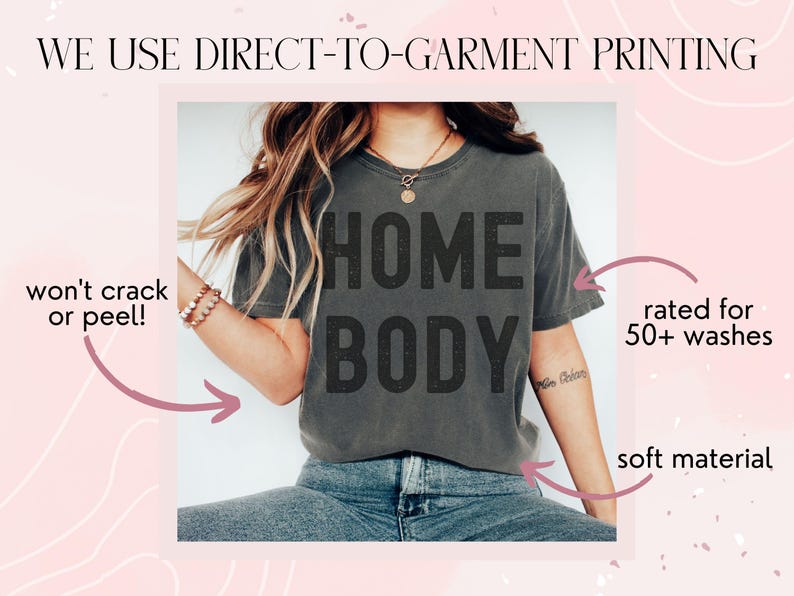 May include: A gray t-shirt with the text "HOME BODY" printed on it. The shirt is being worn by a person and is shown against a pink background with white swirls. The text "WE USE DIRECT-TO-GARMENT PRINTING" is at the top of the image. The text "won't crack or peel!" is on the left side of the image. The text "rated for 50+ washes" is on the right side of the image. The text "soft material" is at the bottom right of the image.