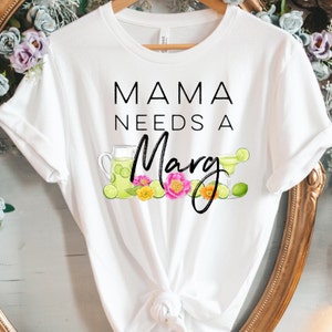 May include: White t-shirt with the text "MAMA NEEDS A Marg" in black script. The design includes illustrations of a pitcher and margarita glass with lime slices, and colorful flowers. A casual, comfortable tee.
