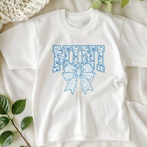 May include: White short-sleeved t-shirt with a blue floral pattern and the word "MINI" in blue with a blue floral bow.