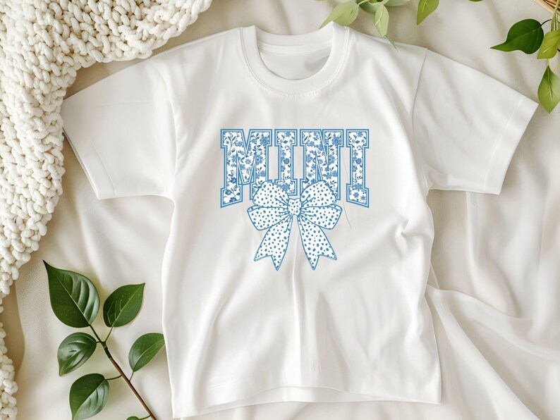 May include: White short-sleeved t-shirt with a blue floral pattern and the word "MINI" in blue with a blue floral bow.