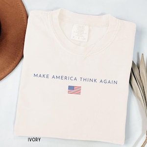 May include: Ivory-colored t-shirt with the text "MAKE AMERICA THINK AGAIN" in navy blue, along with a small American flag. A brown hat and dried leaves are in the background.
