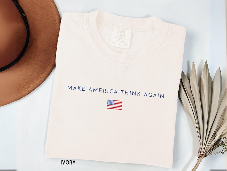 May include: Ivory-colored t-shirt with the text "MAKE AMERICA THINK AGAIN" in navy blue, along with a small American flag. A brown hat and dried leaves are in the background.