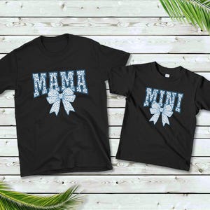 May include: Two black t-shirts with a white and blue bow design. The larger shirt says "MAMA" and the smaller shirt says "MINI".