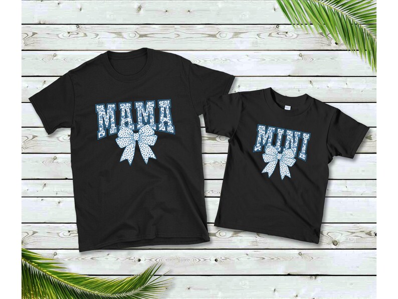 May include: Two black t-shirts with a white and blue bow design. The larger shirt says "MAMA" and the smaller shirt says "MINI".