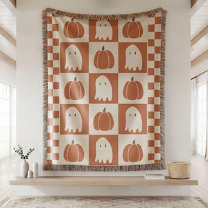 May include: A Halloween-themed throw blanket with a repeating pattern of pumpkins and ghosts in a grid. The blanket is primarily cream and orange, with a checkered border and fringed edges. The design is simple and graphic, perfect for seasonal decor.