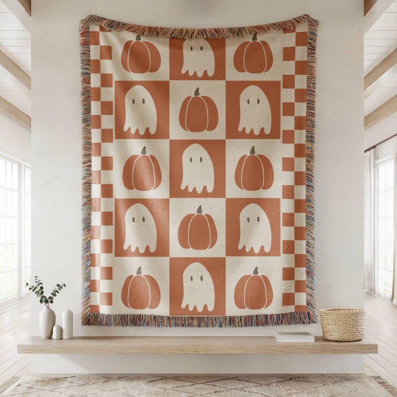 May include: A Halloween-themed throw blanket with a repeating pattern of pumpkins and ghosts in a grid. The blanket is primarily cream and orange, with a checkered border and fringed edges. The design is simple and graphic, perfect for seasonal decor.