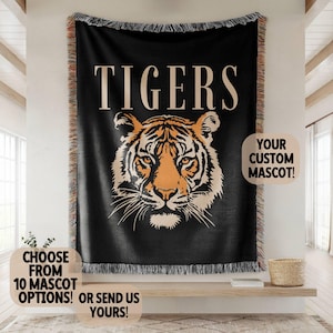 May include: A black woven throw blanket featuring a large tiger face graphic in orange and cream tones. The word TIGERS is printed in large cream letters above the tiger. The blanket has fringed edges and includes text about custom mascot options.