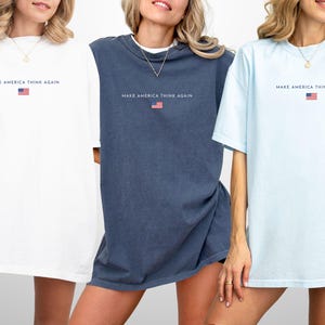 May include: Three oversized t-shirts in white, navy blue, and light blue. Each shirt has the text "MAKE AMERICA THINK AGAIN" above a small American flag graphic. The shirts have short sleeves and a relaxed fit.