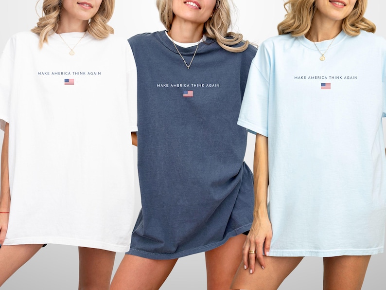May include: Three oversized t-shirts in white, navy blue, and light blue. Each shirt has the text "MAKE AMERICA THINK AGAIN" above a small American flag graphic. The shirts have short sleeves and a relaxed fit.