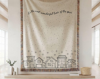 May include: Cream-colored woven throw blanket with black line art of houses and stars. The blanket has a fringed edge with multiple colors and the text "It's the most wonderful time of the year".