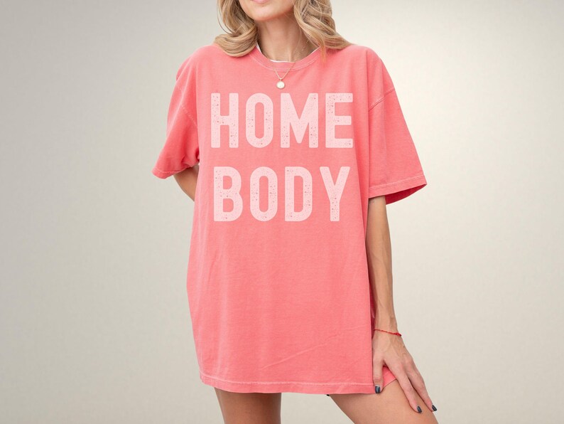 May include: A coral pink oversized t-shirt with the text "HOME BODY" printed in white.