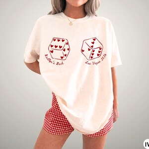 May include: Ivory-colored t-shirt with two dice graphics, each with heart-shaped pips. The text "Kaitlyn's Bach" and "Las Vegas 2026" are printed in red. The model is wearing red and white checkered shorts.