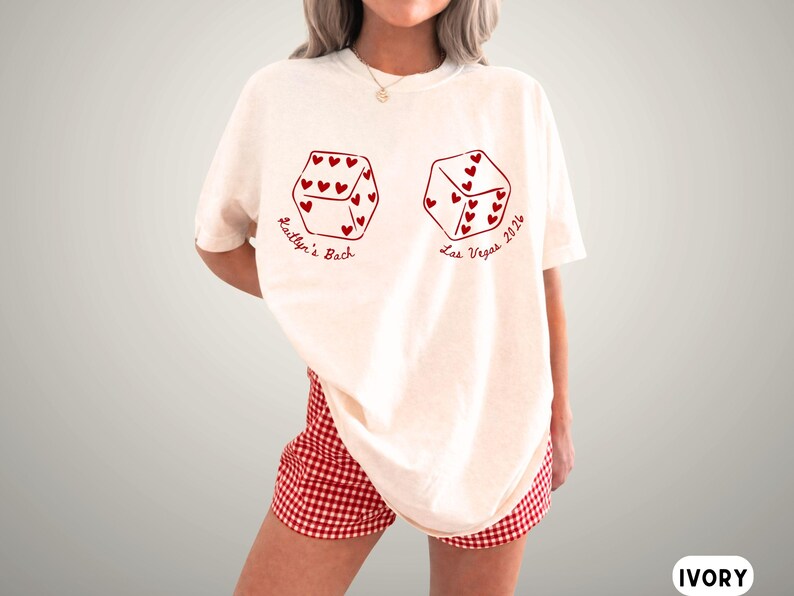 May include: Ivory-colored t-shirt with two dice graphics, each with heart-shaped pips. The text "Kaitlyn's Bach" and "Las Vegas 2026" are printed in red. The model is wearing red and white checkered shorts.