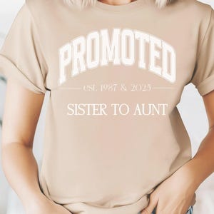 May include: A beige t-shirt with rolled sleeves. The shirt features the text "PROMOTED" in a white, arched font, with "est. 1987 & 2025" below it. Underneath, the text reads "SISTER TO AUNT".