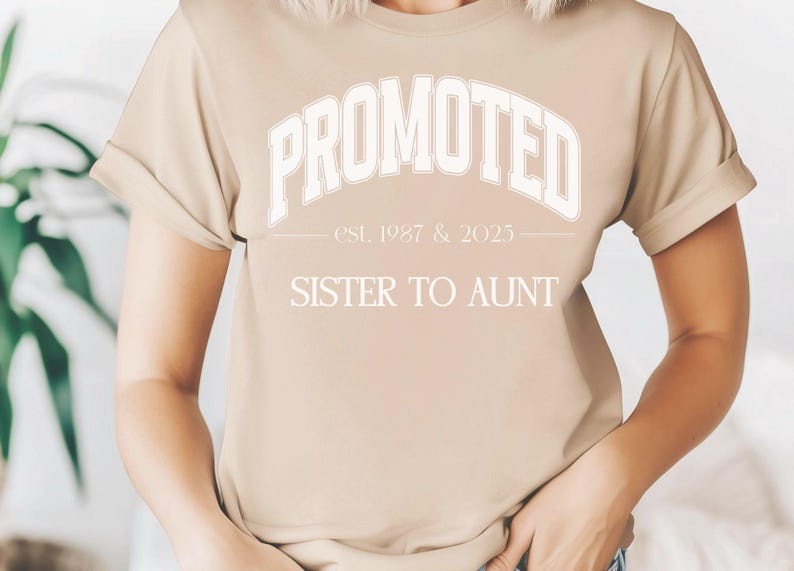 May include: A beige t-shirt with rolled sleeves. The shirt features the text "PROMOTED" in a white, arched font, with "est. 1987 & 2025" below it. Underneath, the text reads "SISTER TO AUNT".