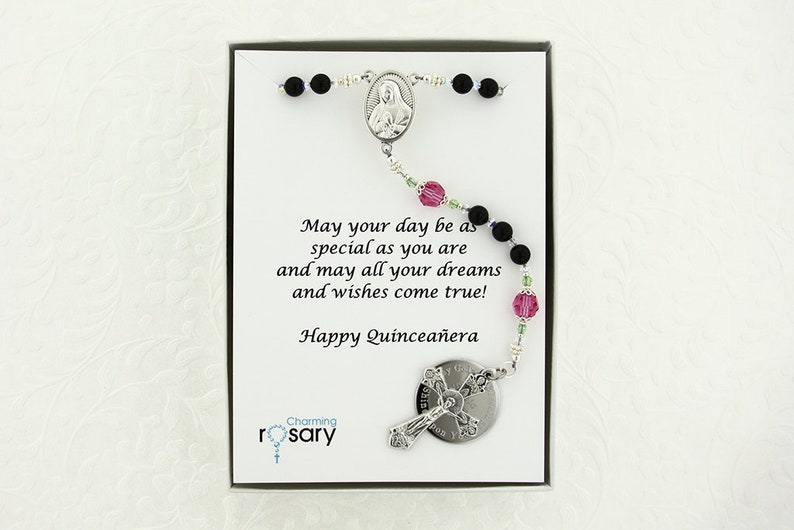 May include: A black and silver rosary with pink and green beads in a white box. The box has a message that reads "May your day be as special as you are and may all your dreams and wishes come true! Happy Quincea&ntilde;era". The box also has the words "Charming rosary" printed on it.